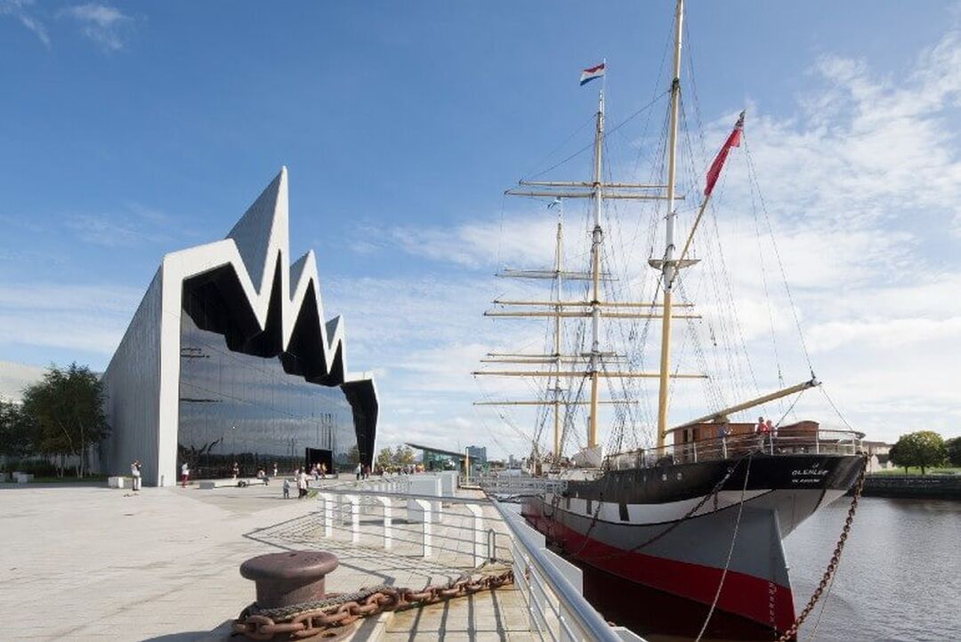 The Tall Ship sits berthed on the River Clyde beside the Riverside Museum. The silver modern building has a wave-like designed roof which spikes into a blue sky.