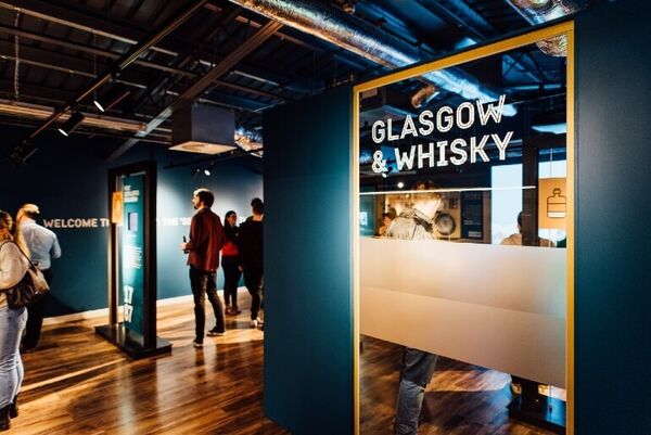 People look around a visitor centre which is modern in style with dark blue walls and exposed ceiling piping. A sign reads 'Glasgow & Whisky'.