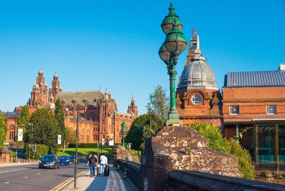 Looking down a main road, the domed roof of Kelvinhall is to one side with Kelvingrove Art Gallery and Museum on the other side. Both are brown sandstone buildings that are Victorian in style.