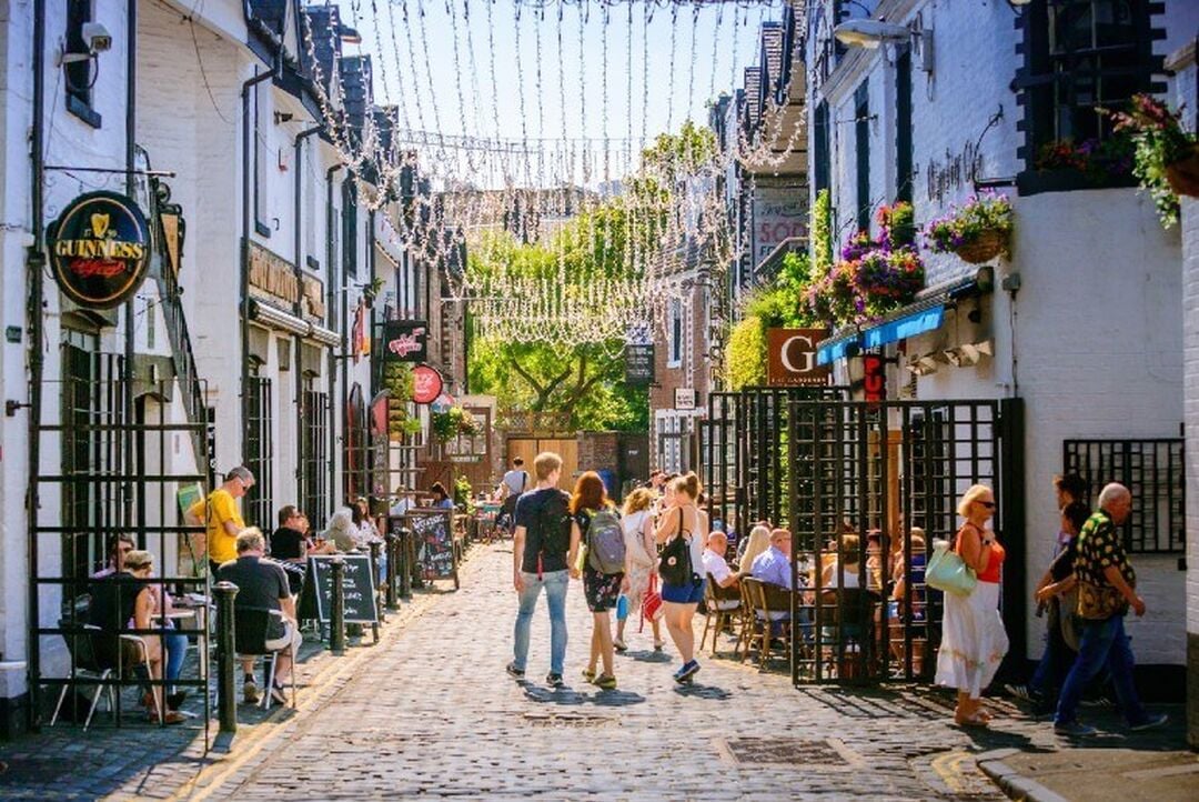 People walk down a cobbled lane with quirky buildings either side, with hanging baskets of flowers and fairy lights overhead.