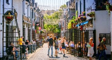 People walk down a cobbled lane with quirky buildings either side, with hanging baskets of flowers and fairy lights overhead.