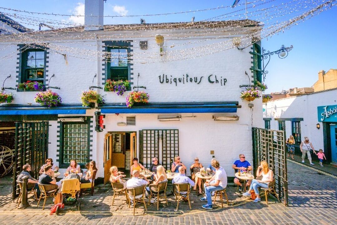 People sit outside on a sunny day around tables in front of a white building with the sign 'Ubiquitous Chip' on the side.