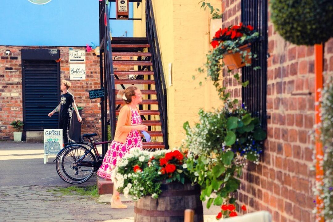 A person in a summer's dress walks between two have exposed bricks and colourful walls. Flower pots and hanging baskets sit outside the buildings.