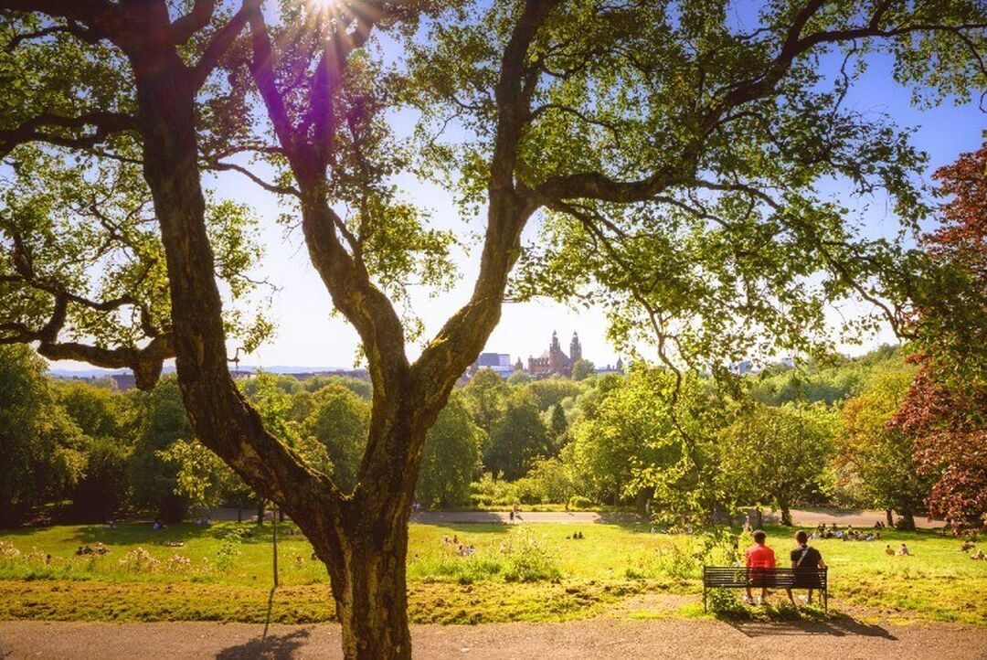 Two people sit on a bench looking out on a green tree-lined view. The turrets of Kelvingrove Art Gallery and Museum can be seen in the distance between trees.