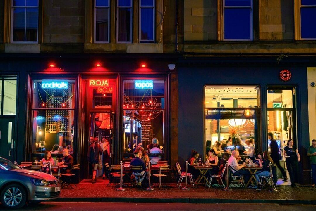 A night-time scene sees people dining outside of cafes and restaurants in front of a road. The red, and yellow lighting from inside the restaurants shines out onto the street.