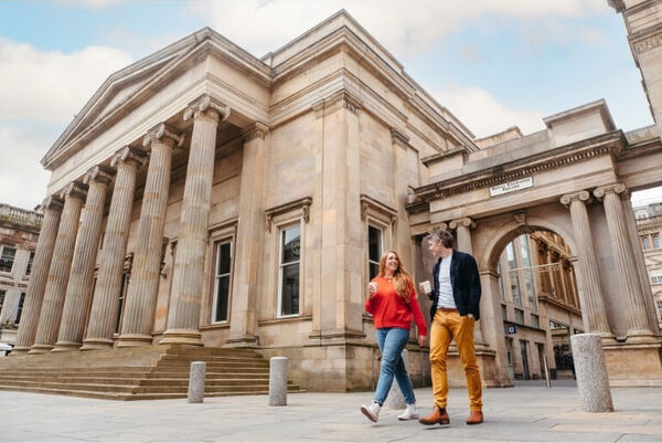 Two people wearing bright clothing holding coffees as they walk past blonde sandstone buildings in Royal Exchange Square.