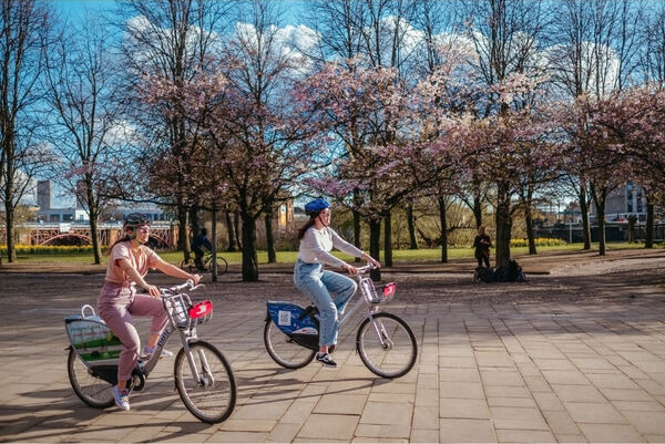 Two people riding Nextbikes with cherry blossom trees in the background