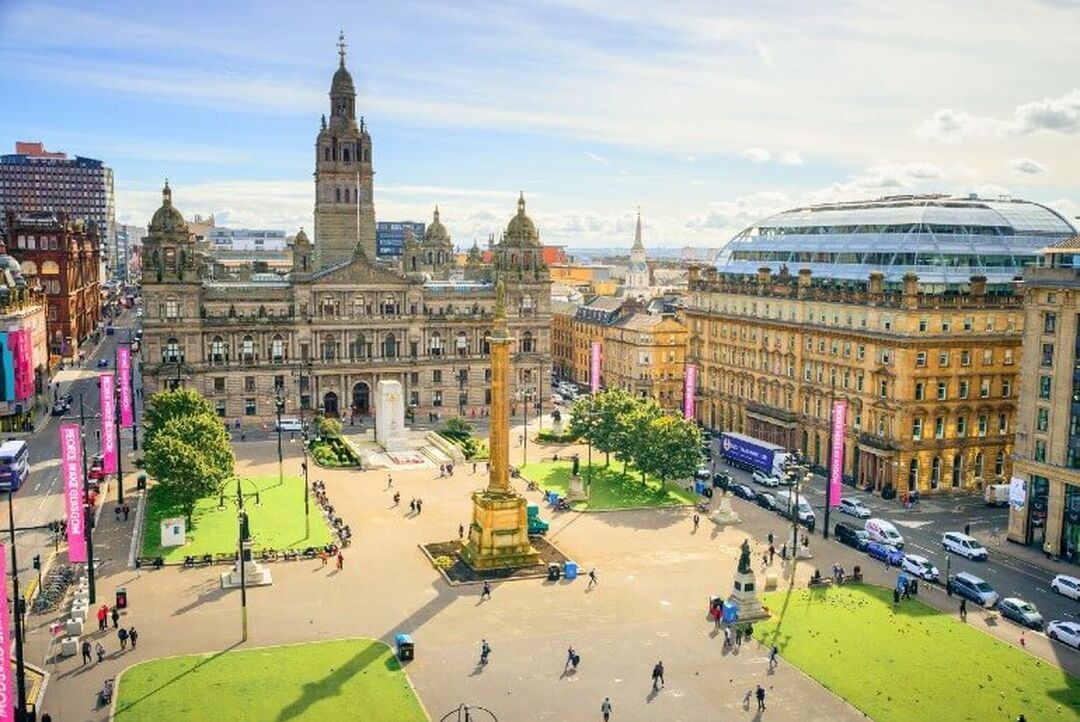 Victorian buildings, including the City Chambers surround the outside of George Square. People are walking through the square which has grass squares and a monument in the centre.