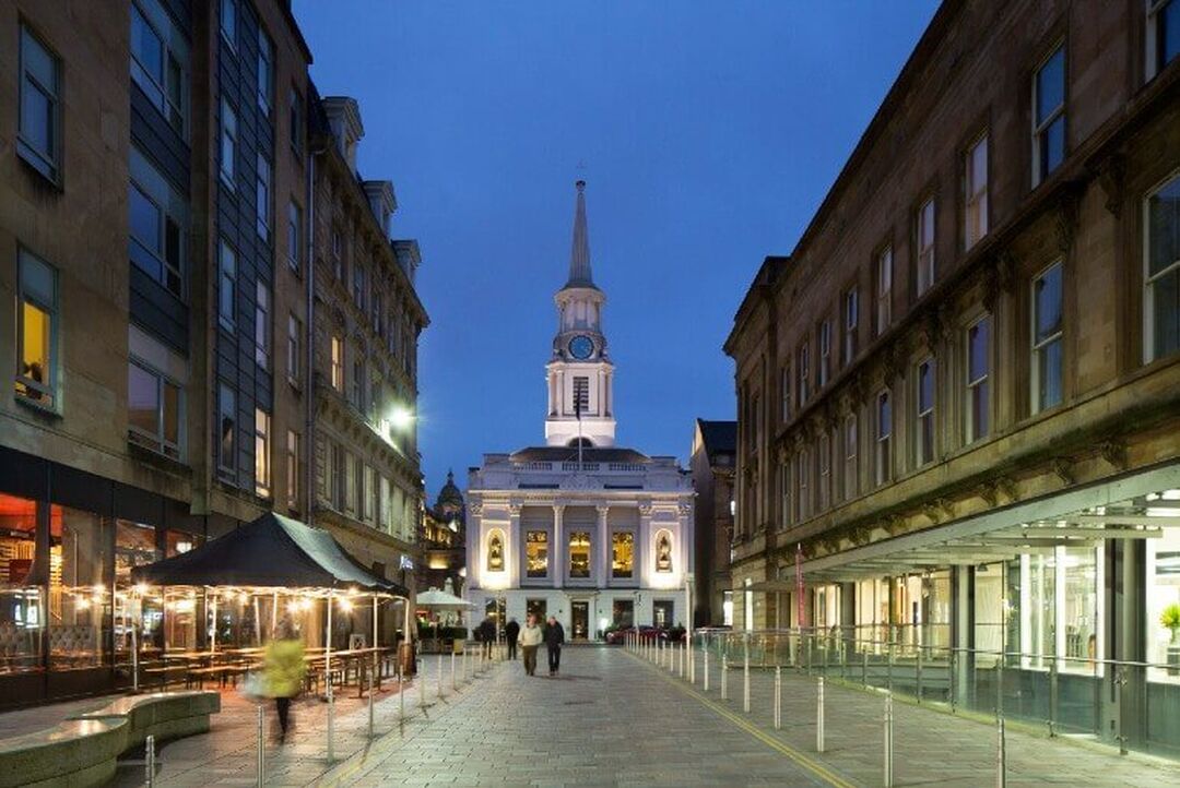 A white church is lit up against the evening sky. It sits at the centre of a cobbled pedestrianised street with well-kept buildings on either side.
