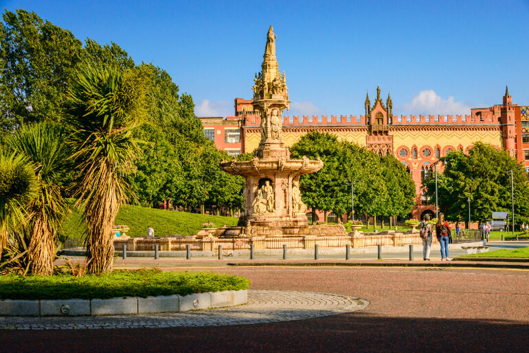 Large terracotta water fountain with green trees, featuring a red and patterned brick building