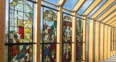 A glass wall and ceiling is lined with a series of wooden beams. 4 of the glass panels have full length stained glass designs.