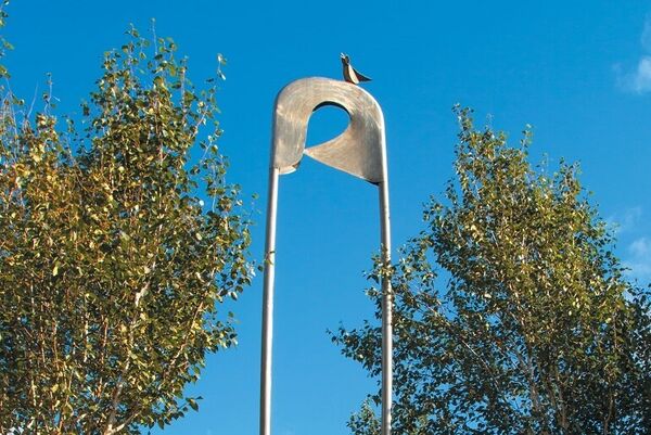 A large metal nappy pin sculpture with a bird atop sits between trees.