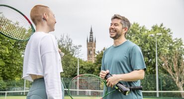2 people talking on a tennis pitch, holding tennis rackets, with a grand building and greenery in the background.